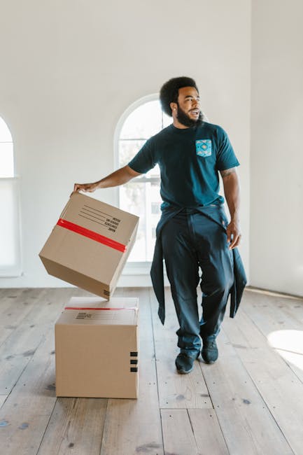 A man with dark curly hair and a beard, dressed in a navy blue t-shirt and dark trousers, is inside a bright, unfurnished room with white walls and wooden flooring. He is holding a medium-sized cardboard box with red tape on top, preparing for a home relocation. The box appears to contain household items and has printed labels. In front of him, on the floor, is another sealed cardboard box. The room has large, arched windows allowing natural light to illuminate the space. The scene suggests a furniture transport or packing process as part of a professional moving service, with the man actively engaged in loading or packing during a house move. The lighting emphasizes a clean, minimalistic environment typical of a property awaiting furniture and belongings to be moved in or out, consistent with services provided by Movers Tufnell Park during house removals.