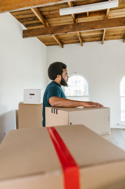 A man with dark curly hair and a beard, dressed in a teal shirt, standing inside a well-lit room with white walls and a wooden ceiling, is preparing for a home relocation. He is positioned behind several large cardboard boxes, some sealed with red packing tape, which are stacked on a smooth wooden floor. The boxes display minimal markings, indicating they are packed for moving. In the background, there are two arched windows allowing natural light to illuminate the space, along with a partially visible open doorway. The overall scene captures the process of packing and preparing for furniture transport, with the man likely involved in the loading process. The room appears to be in the early stages of a house removal, with no furniture or appliances visible, emphasizing the focus on packing materials and the moving logistics managed by Movers Tufnell Park, specialists in removals and home relocation services.